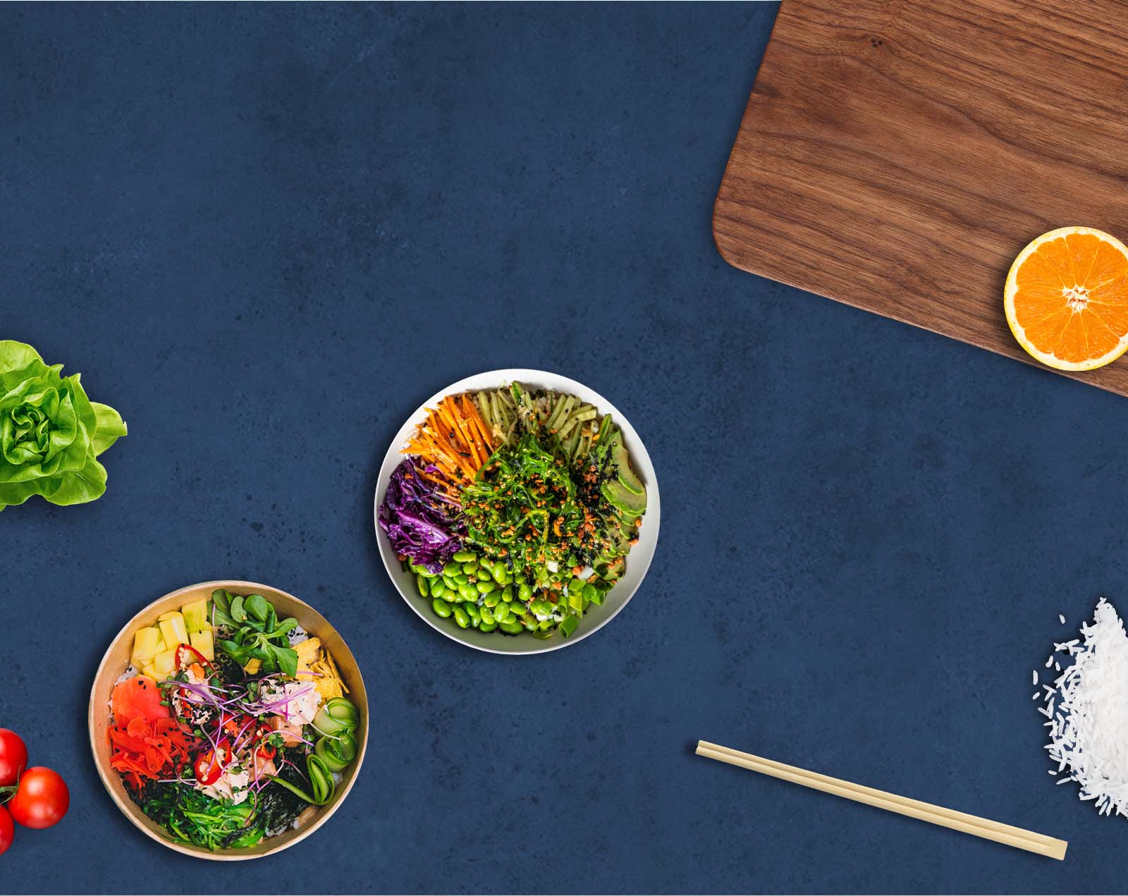 Bols de poké bowl vus de haut sur un plan de travail bleu nuit, entourés de légumes frais, riz et baguettes